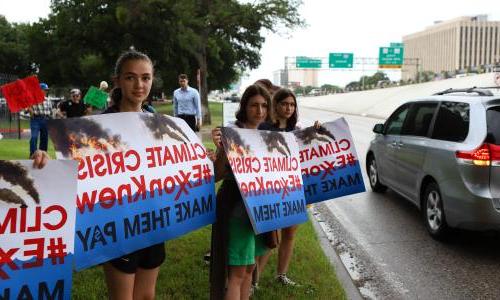 Young girls on the side of the road hold signs that read "Climate Crisis, #ExxonKnew Make Them pay."